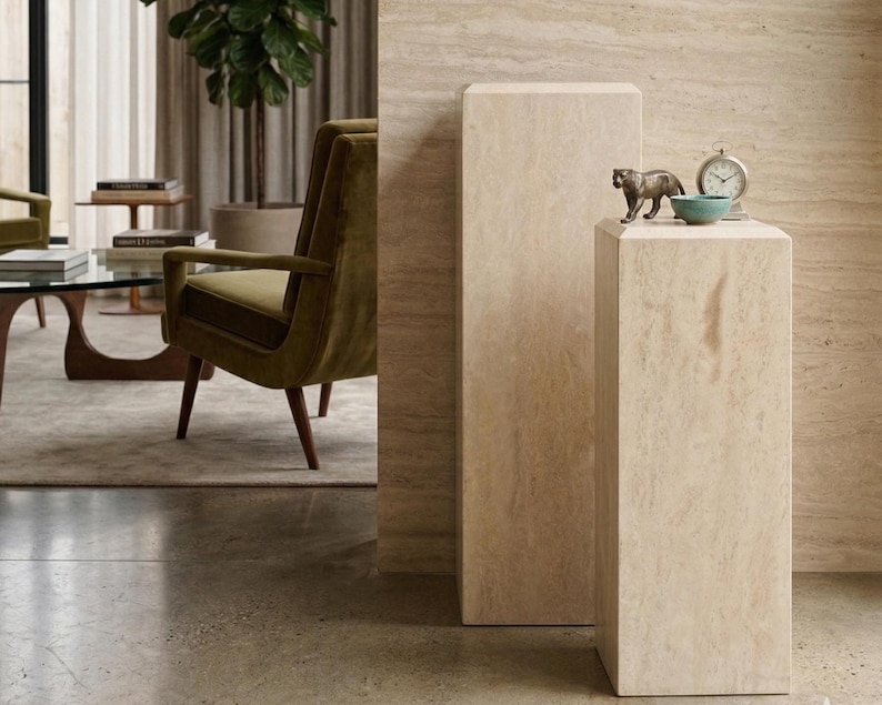 May include: Two rectangular beige travertine stone pedestals of varying heights. The shorter pedestal holds a small bronze-colored tiger figurine, a small teal bowl, and a silver-toned analog clock with a white face and black hands.