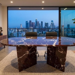 May include: An oval-shaped conference table featuring a deep reddish-brown marble surface with prominent white veining, supported by two matching marble pedestal bases, positioned in front of a large window overlooking a city skyline at dusk.