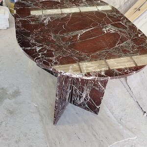 May include: An oval dining table crafted from deep red marble with prominent white veining. The table features a polished, reflective surface and is supported by a central, cross-shaped pedestal base made from the same matching red marble material.