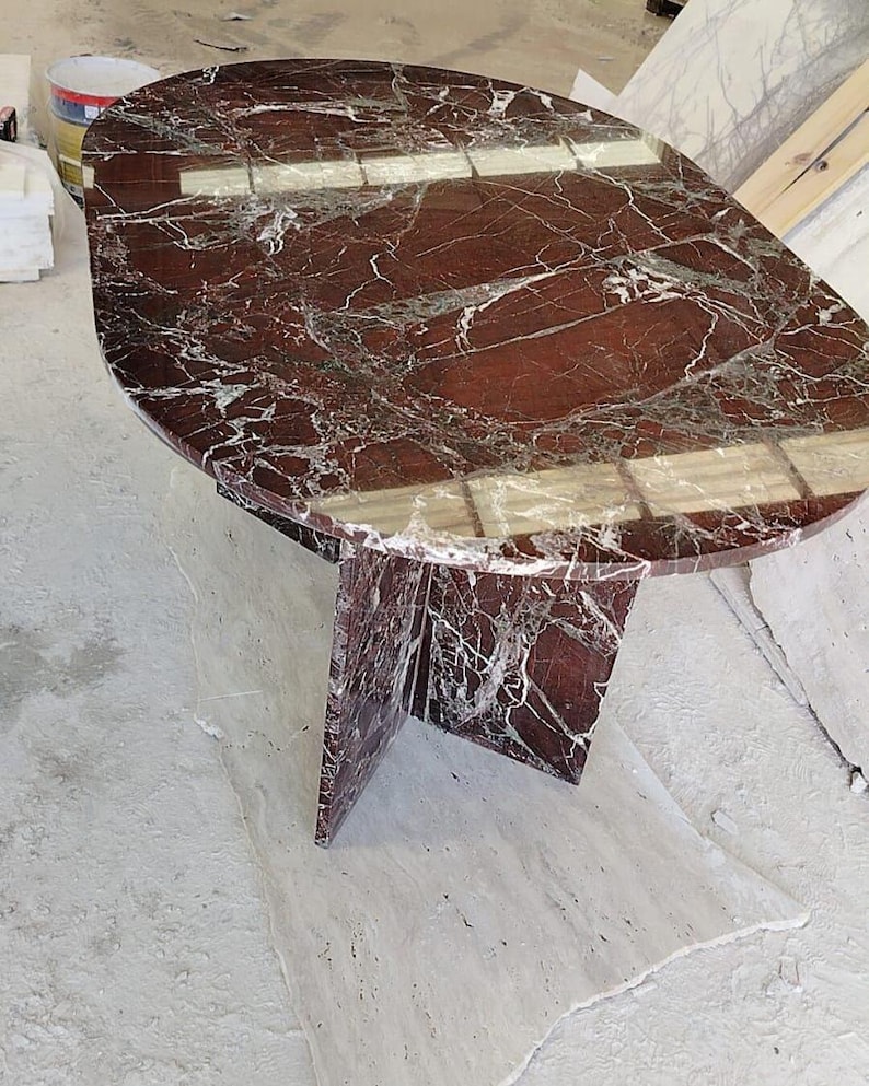 May include: An oval dining table crafted from deep red marble with prominent white veining. The table features a polished, reflective surface and is supported by a central, cross-shaped pedestal base made from the same matching red marble material.