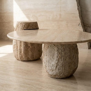 May include: A beige stone coffee table with a round top and two cylindrical bases. A matching stone stool is behind the table. The furniture has a natural, textured appearance, suitable for a modern living space.