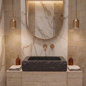 Pu&ograve; includere: Interni di un bagno moderno con un lavabo in pietra grigio scuro, rubinetti dorati e un grande specchio ovale. Due bottiglie di vetro ambrato sono sul bancone, con due lampade a sospensione dorate sopra. Le pareti sono un mix di marmo e piastrelle quadrate.