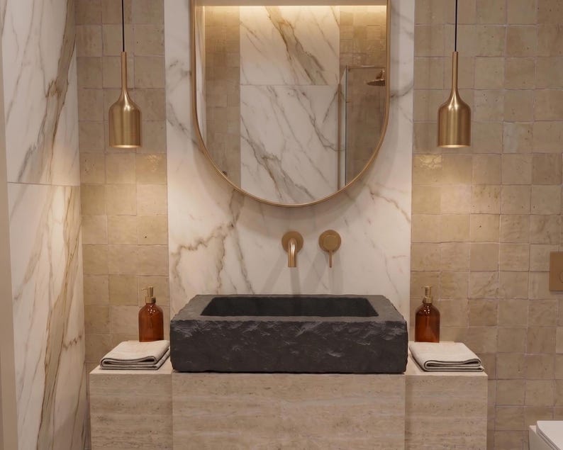 Pu&ograve; includere: Interni di un bagno moderno con un lavabo in pietra grigio scuro, rubinetti dorati e un grande specchio ovale. Due bottiglie di vetro ambrato sono sul bancone, con due lampade a sospensione dorate sopra. Le pareti sono un mix di marmo e piastrelle quadrate.