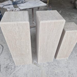 May include: Three rectangular stone pedestals of varying heights arranged in a row. Each pedestal is crafted from light beige travertine with a natural, porous, and veined texture, featuring a smooth, flat top surface and clean, straight edges.