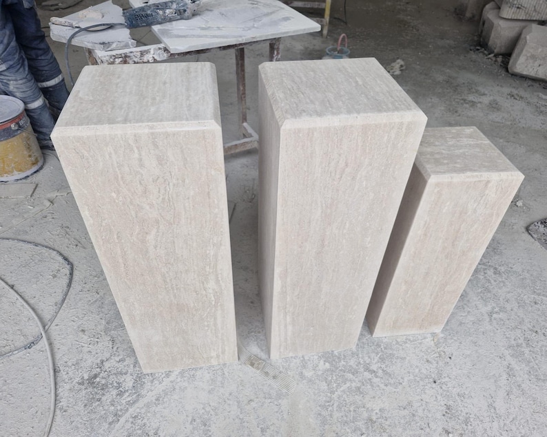 May include: Three rectangular stone pedestals of varying heights arranged in a row. Each pedestal is crafted from light beige travertine with a natural, porous, and veined texture, featuring a smooth, flat top surface and clean, straight edges.