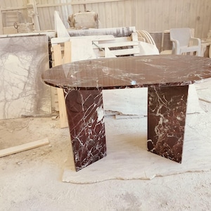 May include: An oval dining table crafted from deep red marble with prominent white veining. The table features a smooth, polished oval top supported by two thick, rectangular slab legs made of the same matching red and white marble material.
