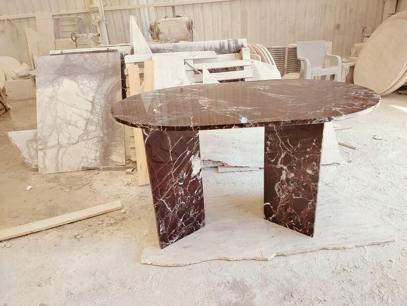 May include: An oval dining table crafted from deep red marble with prominent white veining. The table features a smooth, polished oval top supported by two thick, rectangular slab legs made of the same matching red and white marble material.