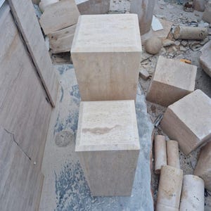 May include: Two square, beige stone pedestals with a natural, porous texture. The pedestals are positioned vertically, one behind the other, showcasing a light tan color with subtle variations in the stone grain and a smooth, flat top surface.
