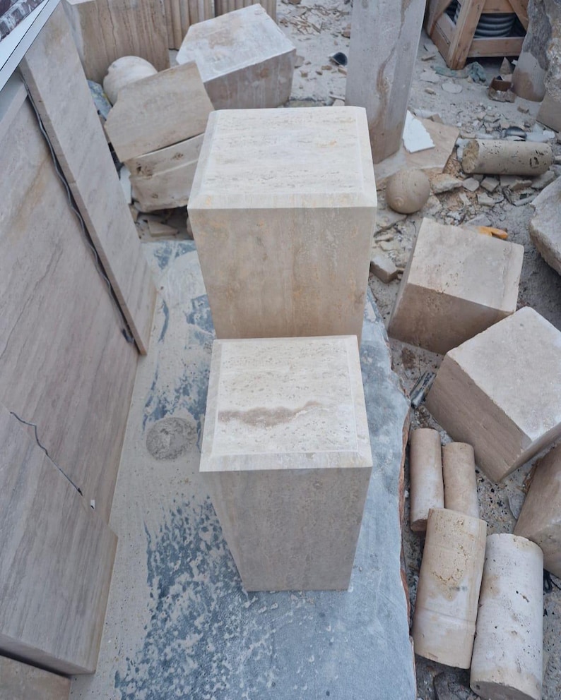 May include: Two square, beige stone pedestals with a natural, porous texture. The pedestals are positioned vertically, one behind the other, showcasing a light tan color with subtle variations in the stone grain and a smooth, flat top surface.