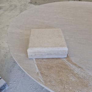 May include: A square beige stone block with a smooth top surface and beveled edges, featuring natural horizontal veining and a porous texture, resting on a large, circular, light-colored stone slab with a similar natural finish.