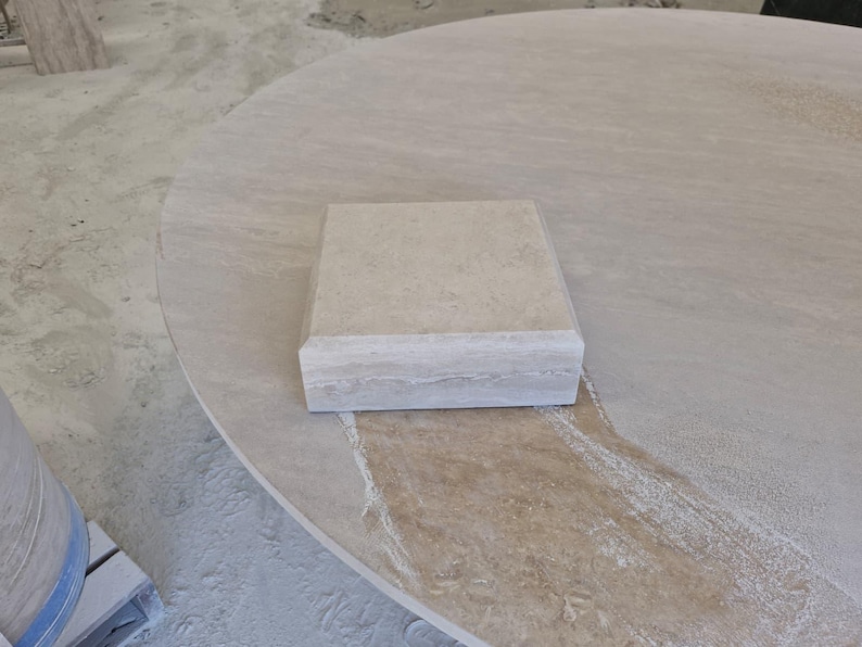 May include: A square beige stone block with a smooth top surface and beveled edges, featuring natural horizontal veining and a porous texture, resting on a large, circular, light-colored stone slab with a similar natural finish.