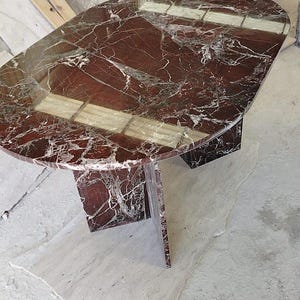 May include: An oval dining table crafted from deep red marble with prominent white veining. The table features a polished, reflective surface and is supported by a central pedestal base made of the same red marble material.