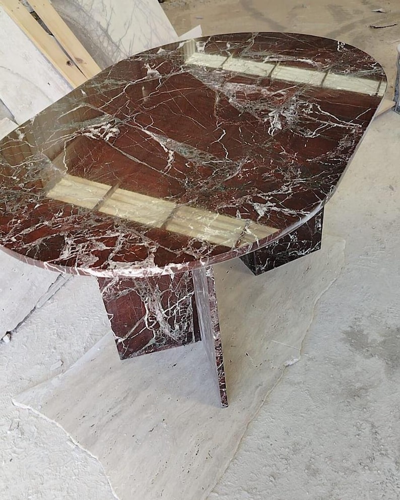 May include: An oval dining table crafted from deep red marble with prominent white veining. The table features a polished, reflective surface and is supported by a central pedestal base made of the same red marble material.