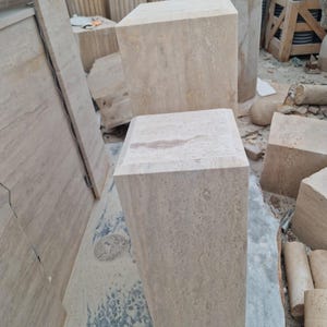 May include: Two rectangular stone pedestals made of light beige travertine with a natural porous texture. The pedestals feature flat, square tops and smooth vertical sides, showcasing the organic veining and earthy tones of the natural stone material.