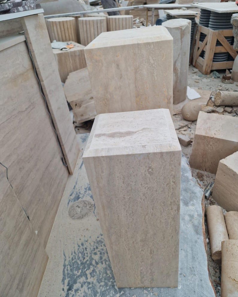 May include: Two rectangular stone pedestals made of light beige travertine with a natural porous texture. The pedestals feature flat, square tops and smooth vertical sides, showcasing the organic veining and earthy tones of the natural stone material.