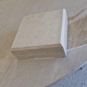 May include: A square, light beige stone block with a smooth, flat top surface and a rounded, bullnose-style edge profile. The stone features a natural, subtle mottled texture with faint horizontal veining visible along the side thickness.