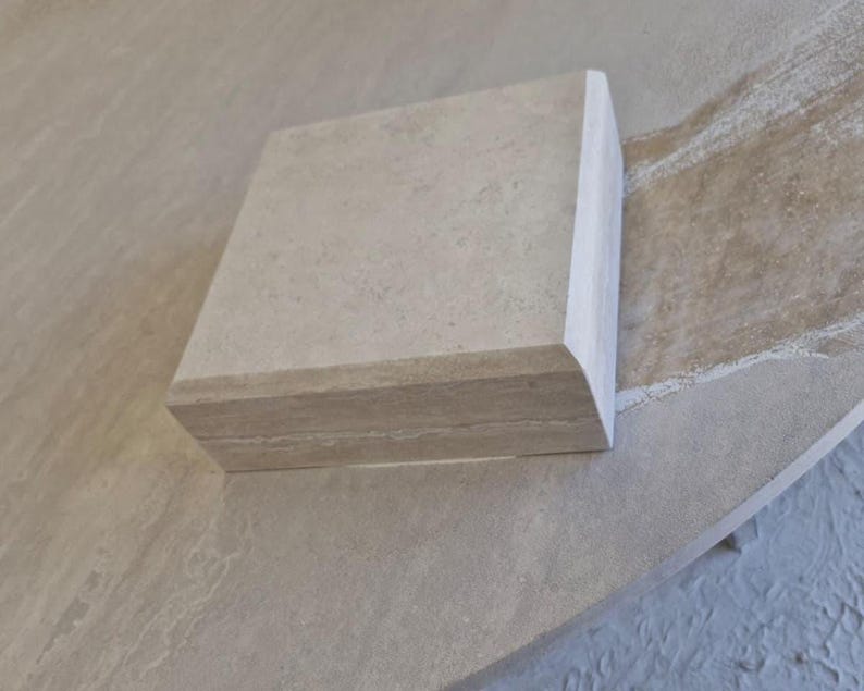May include: A square, light beige stone block with a smooth, flat top surface and a rounded, bullnose-style edge profile. The stone features a natural, subtle mottled texture with faint horizontal veining visible along the side thickness.