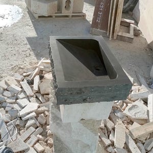 Pu&ograve; includere: Un lavandino in pietra grigio scuro con un bacino rettangolare e un bordo grezzo. Il lavandino &egrave; montato su un piedistallo in pietra di colore chiaro. Lo sfondo mostra materiali da costruzione e detriti.