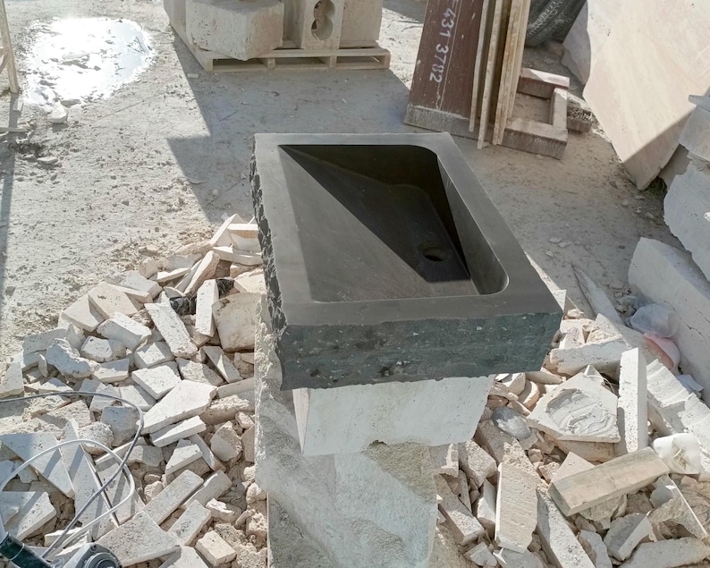 Pu&ograve; includere: Un lavandino in pietra grigio scuro con un bacino rettangolare e un bordo grezzo. Il lavandino &egrave; montato su un piedistallo in pietra di colore chiaro. Lo sfondo mostra materiali da costruzione e detriti.