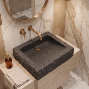 Pu&ograve; includere: Un lavabo moderno in bagno in pietra grigio scuro. Il lavabo &egrave; rettangolare con un bordo ruvido e naturale. Un rubinetto dorato e accessori abbinati sono montati a parete. Un dosatore di sapone marrone e un asciugamano piegato sono sul bancone.