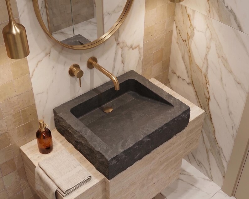 Pu&ograve; includere: Un lavabo moderno in bagno in pietra grigio scuro. Il lavabo &egrave; rettangolare con un bordo ruvido e naturale. Un rubinetto dorato e accessori abbinati sono montati a parete. Un dosatore di sapone marrone e un asciugamano piegato sono sul bancone.