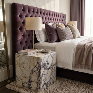 May include: A bedroom scene featuring a tall, deep purple velvet tufted headboard and a white marble cube nightstand with dark veining. A gold lamp with a beige shade, a book titled The Soul of Style, and a small bowl sit on the marble surface.