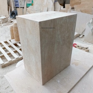 May include: A rectangular block made of beige travertine stone with a natural porous texture. The front face features the text NIZA DEKARASYON printed in black. The object has sharp, clean edges and a solid, monolithic appearance with light tan veining.