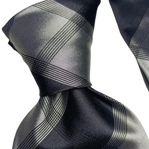 Può includere: Primo piano di una cravatta di seta con motivo a quadri. La cravatta presenta una combinazione di toni neri e grigi, con un design a griglia diagonale. La cravatta è annodata, mostrando la sua consistenza e lucentezza.