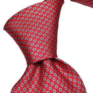 Könnte beinhalten: Eine rote Krawatte mit einem sich wiederholenden Muster aus kleinen weißen und blauen blumenartigen geometrischen Formen. Die Krawatte ist gebunden und zeigt die strukturierte Stoffoberfläche sowie die gleichmäßige Verteilung der Motive.