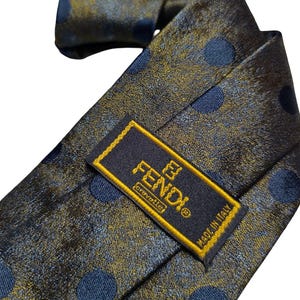 May include: A Fendi tie with a navy blue and gold polka dot pattern. The tie has a label that reads "FENDI cravatte MADE IN ITALY". The tie is made of a shiny material.
