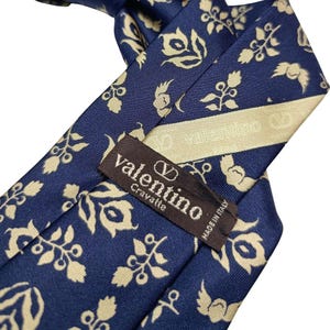 Könnte beinhalten: Eine marineblaue Krawatte mit einem sich wiederholenden cremefarbenen Blumenmuster. Auf der Rückseite ist ein braunes Etikett aufgenäht, das den Text "valentino Cravatte" mit einem Logo darüber und "MADE IN ITALY" vertikal rechts zeigt.
