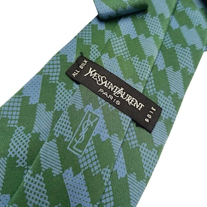 May include: A green and blue patterned silk tie. The tie features a repeating geometric design in shades of green and blue. The label reads "Yves Saint Laurent Paris" with the brand's logo.