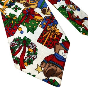 Puede incluir: Una corbata blanca con temática navideña con un patrón repetido de osos de peluche, regalos envueltos, árboles de Navidad y coronas. La corbata tiene una forma clásica y está hecha de una tela suave. El diseño incluye acentos rojos, verdes, azules y amarillos.