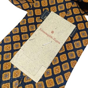Könnte beinhalten: Eine marineblaue Krawatte mit einem sich wiederholenden Muster aus goldenen und blauen Quadraten. Das Etikett lautet "Ermenegildo Zegna". Die Krawatte ist aus glattem Stoff, wahrscheinlich Seide.
