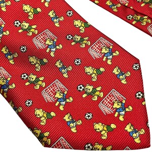 Puede incluir: Una corbata roja con un patrón repetitivo de osos de peluche amarillos jugando al fútbol. Los osos aparecen en varias posturas, algunos con camisetas azules o verdes, junto a balones de fútbol blancos y porterías de color rojo y blanco.