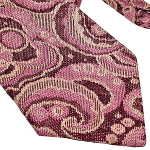 Pode incluir: Uma gravata com um padrão paisley tecido em tons de bordeaux profundo, magenta e bege claro. O tecido tem uma aparência texturizada com motivos intrincados em espiral que cobrem toda a superfície da ponta da gravata.