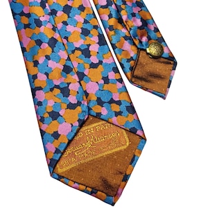 Può includere: Una cravatta di seta colorata con un motivo ripetuto di forme blu, arancioni e rosa su sfondo blu navy. La cravatta ha un retro marrone e un bottone dorato. L'etichetta recita "Made in Italy".