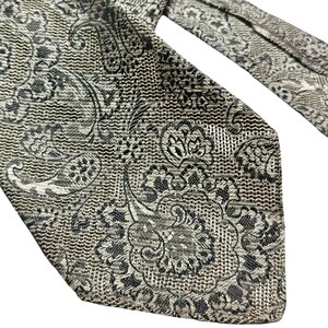 Pode incluir: Uma gravata cinza e preta com padrão paisley. A gravata está amarrada, mostrando os intrincados desenhos florais e curvos. O tecido tem uma aparência texturizada, com uma mistura de tons cinza claro e escuro.