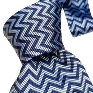 May include: A patterned necktie in shades of blue and white, featuring a zig-zag design. The tie is knotted, highlighting the intricate pattern and colour variations. The design incorporates various shades of blue and white, creating a stylish accessory.