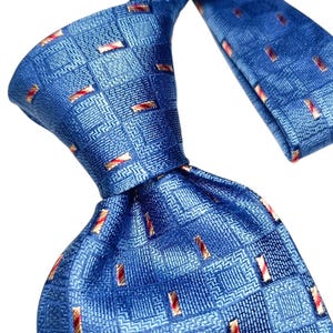 Może przedstawiać: Niebieski jedwabny krawat z powtarzającym się wzorem małych czerwonych i złotych prostokątów. Krawat jest zawiązany, pokazując przód i tył materiału. Tło jest białe.