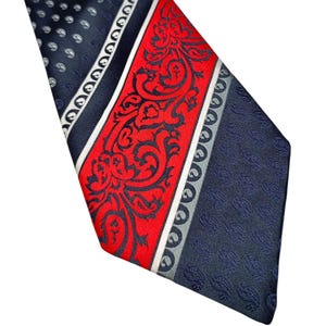 Peut inclure: Une cravate bleu marine avec un motif rouge et blanc. La cravate présente une bande centrale rouge avec un motif orné, bordée de rayures blanches et grises. Les sections bleu marine ont un petit motif de points blancs.