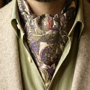 Può includere: Un foulard ascot con una stampa botanica e architettonica dettagliata nei toni del viola, verde e crema. Il tessuto è piegato e infilato nel colletto di una camicia verde chiaro, indossata sotto un blazer beige con motivo a spina di pesce.
