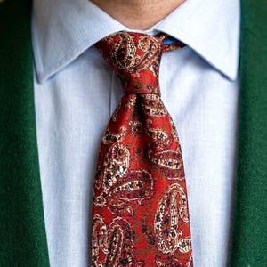 Può includere: Una cravatta rossa con un dettagliato motivo paisley marrone e crema, indossata con una camicia a colletto azzurro e un blazer verde scuro. La cravatta è annodata al centro e presenta una trama tessuta con intricati motivi a vortice.