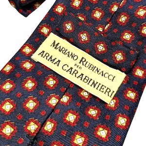 以下が含まれることがあります： 赤と黄色の花の形が繰り返される模様の紺色のネクタイ。「MARIANO RUBINACCI PER ARMA CARABINIERI」と書かれたラベルが付いています。ネクタイは、滑らかな素材、おそらくシルクで作られています。