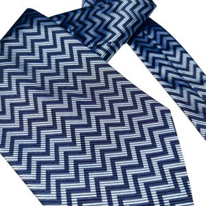 May include: A blue and white patterned tie with a repeating zigzag design. The tie is made of a shiny material and is knotted at the top. The pattern is a mix of dark and light blue with white accents. The tie's wider end is visible.