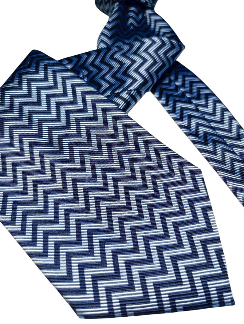 May include: A blue and white patterned tie with a repeating zigzag design. The tie is made of a shiny material and is knotted at the top. The pattern is a mix of dark and light blue with white accents. The tie's wider end is visible.