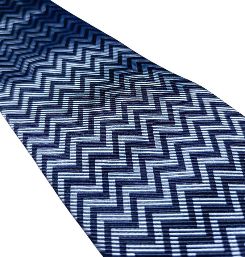 May include: A blue tie with a geometric zigzag pattern. The tie features a repeating design of navy, light blue, and white lines. The fabric has a subtle sheen, and the pattern is consistent across the tie.