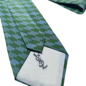 May include: Green and blue patterned tie with the Yves Saint Laurent logo. The tie features a repeating geometric design in shades of green and blue. The back of the tie has a white lining with the YSL logo and "PARIS" printed.
