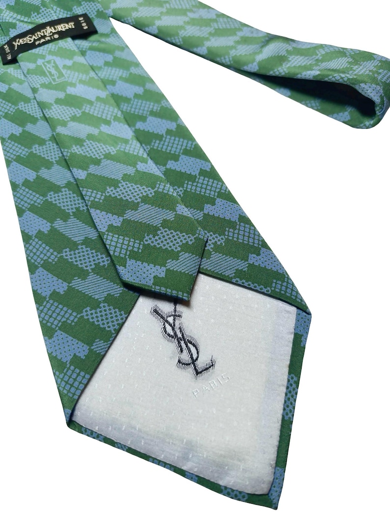 May include: Green and blue patterned tie with the Yves Saint Laurent logo. The tie features a repeating geometric design in shades of green and blue. The back of the tie has a white lining with the YSL logo and "PARIS" printed.