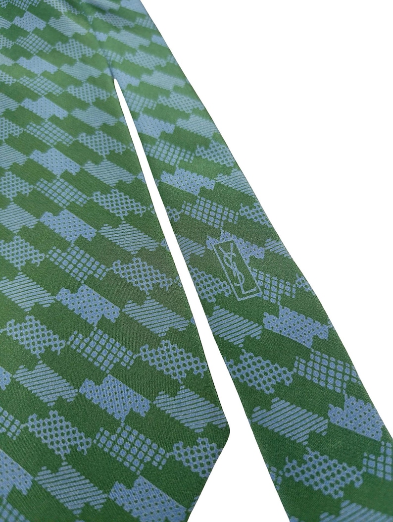May include: Green and blue patterned tie. The tie features a repeating geometric design in shades of green and blue. The YSL logo is visible near the tip of the tie. The tie is made of a smooth fabric.